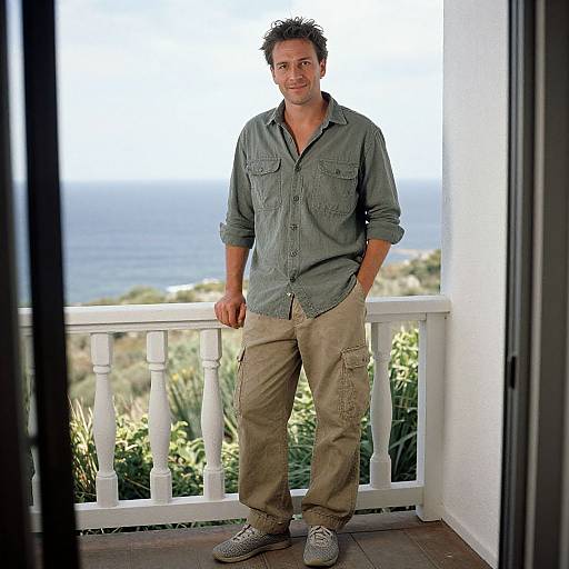 Photograph of a smiling, casually dressed man with short, curly brown hair, wearing a green button-up shirt and beige cargo pants, standing on a