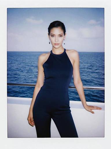 Photograph of a slim, fair-skinned Asian woman with dark hair in a sleek bun, wearing a black halter top and pants, standing against
