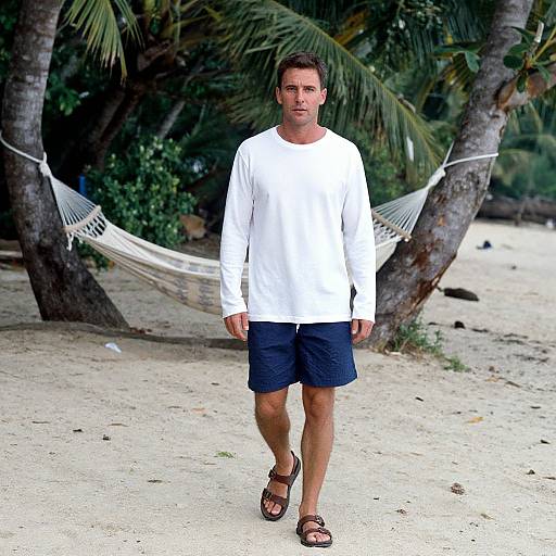 Photograph of a man with short brown hair, wearing a white long-sleeve shirt, navy shorts, and brown sandals, walking on a sandy