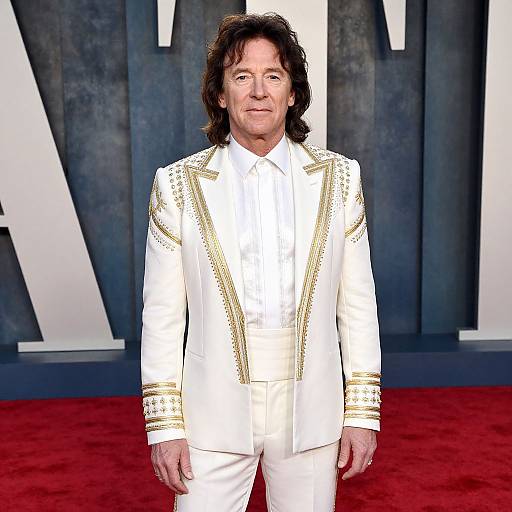 Photograph of middle-aged man with long brown hair, wearing a white, gold-embellished suit, standing on red carpet, large white letter