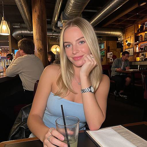 Photograph of a blonde woman with blue eyes, wearing a light blue strapless top, sitting in a dimly lit, industrial-style bar, holding