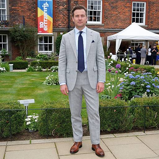 Photograph of a tall, white man in light gray checkered suit, blue tie, brown shoes, standing in front of a brick building with flower