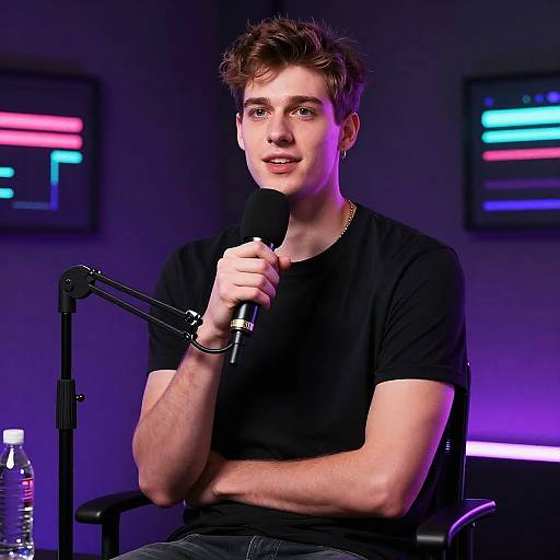 Photograph of a young, fair-skinned man with messy brown hair, wearing a black t-shirt, speaking into a microphone against a dark, neon