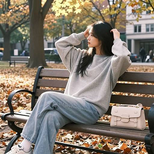 Photograph of a young woman with long black hair, wearing a gray sweatshirt and loose blue jeans, sitting on a park bench, adjusting her hair