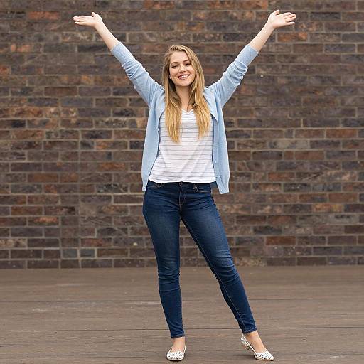 Photograph of a smiling, long-haired blonde woman with arms raised, wearing a light blue cardigan, white striped shirt, dark jeans, and white