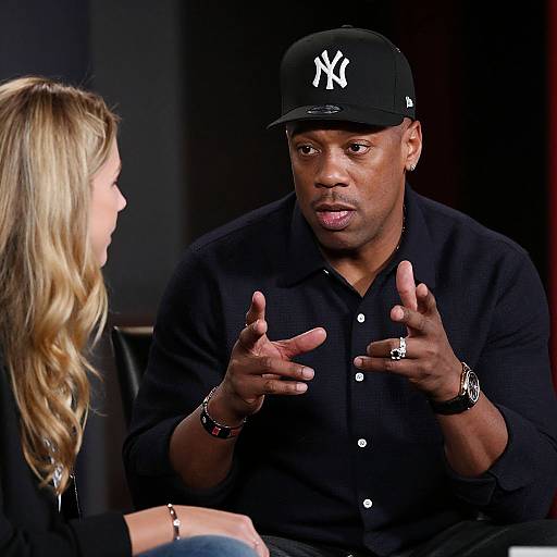 Photograph of a black man in a black NY cap and shirt, gesturing with hands, talking to a blonde woman in a black top. Dim