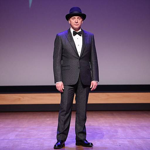 Photograph of a man in a black tuxedo, bow tie, and black top hat, standing on a stage with purple lighting.