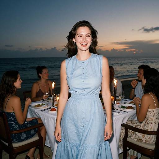 Photograph of a smiling woman in a light blue button-up dress, standing in front of a candlelit dinner table by the sea at sunset.
