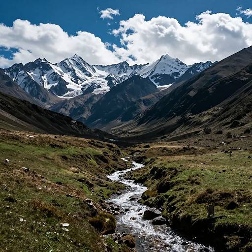 Photograph of a dramatic mountain landscape with snow-capped peaks under a bright blue sky with scattered white clouds. A narrow, flowing river cuts through the