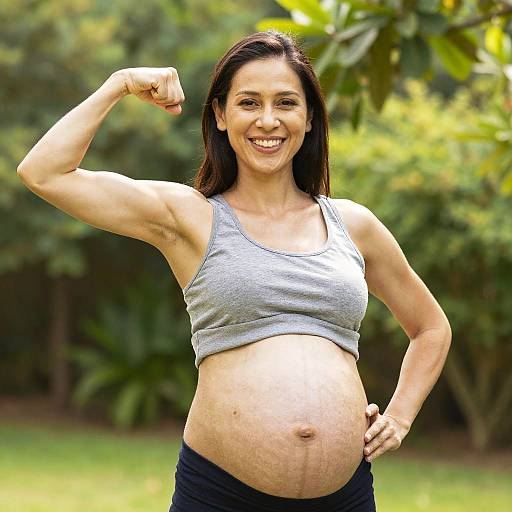 Pregnant woman with long dark hair, smiling, flexing arm in gray sports top, black pants, standing outdoors, green leafy background.