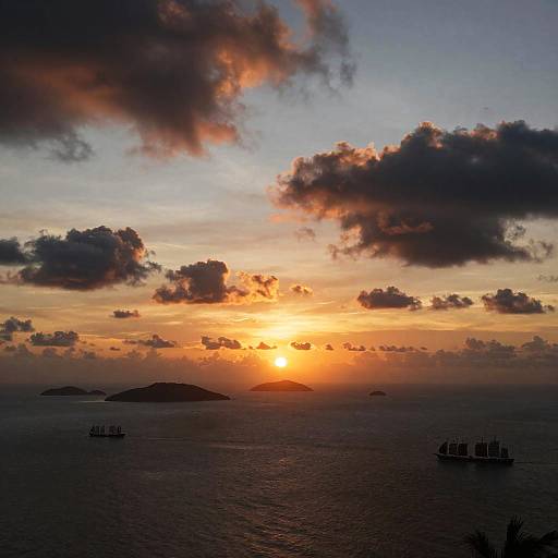 Photograph of a vibrant sunset over a calm ocean, with dark silhouetted clouds, distant islands, and ships in the foreground.