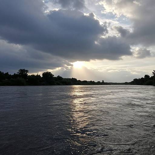 Photograph of a calm, reflective river at sunset with dark, cloudy sky and silhouetted trees on the horizon. Sunlight breaks through clouds