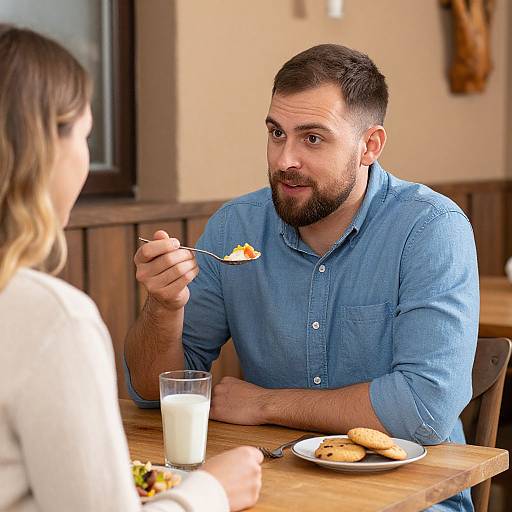 Photograph of a bearded man in a blue shirt, holding a fork with food, sitting at a wooden table with a glass of milk and a