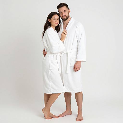 Photograph of a couple in white bathrobes, standing closely, barefoot, against a white background, with the woman's hand on the man
