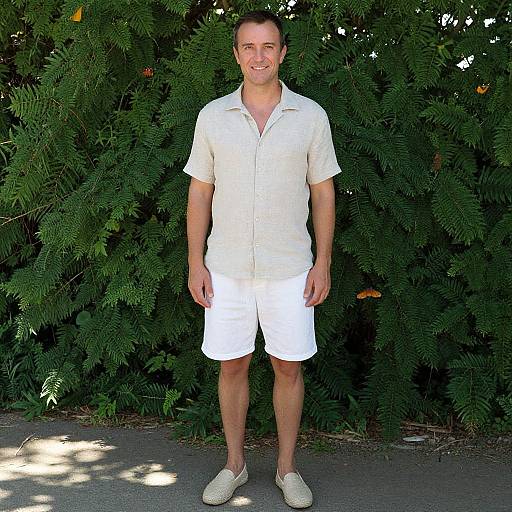 Photograph of a smiling man with short brown hair, wearing a white short-sleeve button-up shirt, white shorts, and white sneakers, standing