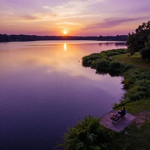 Photograph of a tranquil lake at sunset, with a silhouetted bench on the right, reflecting vibrant orange, pink, and purple skies.