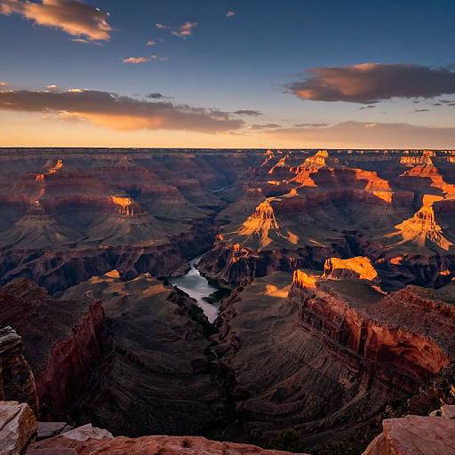 Photograph of the Grand Canyon at sunset, showcasing vibrant orange and red rock formations, deep shadows, a winding river below, and a dramatic sky with