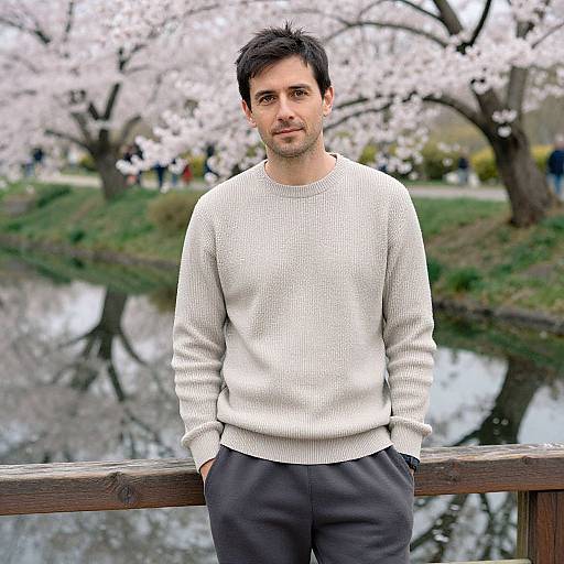 Photograph of a young man with short dark hair, wearing a white knit sweater and black pants, leaning on a wooden railing in front of a cherry
