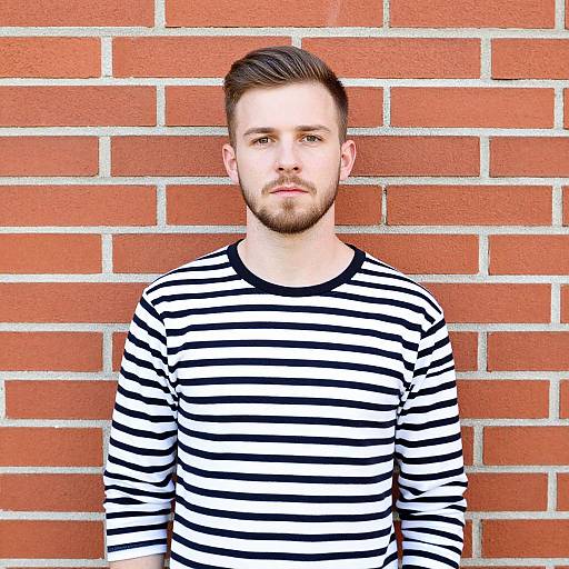 Photograph of a young man with short brown hair and beard, wearing a black-and-white striped long-sleeve shirt, standing against a red brick