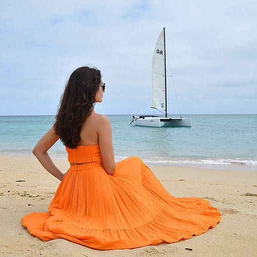 Photograph of a woman with long brown hair in an orange strapless dress, seated on a sandy beach, watching a white sailboat on calm blue