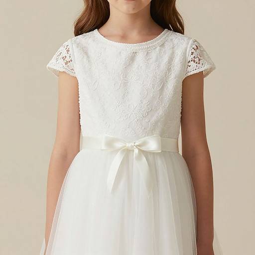 Photograph of a young girl in a white lace short-sleeve dress with a satin bow at the waist, standing against a plain beige background.