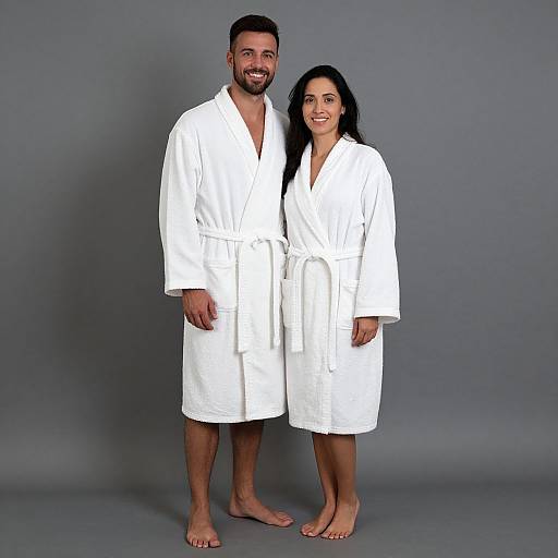 Photograph of a smiling couple in white bathrobes standing barefoot against a gray background, both with dark hair and light skin.