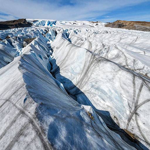 Photograph of a vast, icy glacier with white and blue snow, dark shadows, and a clear blue sky in the background.