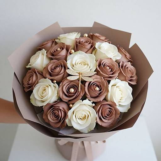 Photograph of a bouquet with alternating white and brown roses, a detailed dragonfly decoration, wrapped in brown paper, held by a hand.