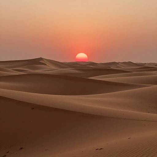 Photograph of a serene desert sunset with a glowing red sun setting behind undulating sand dunes, casting warm orange and red hues across the scene.