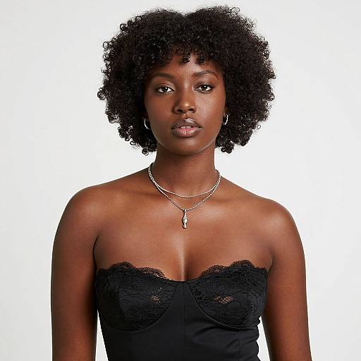 Photograph of a young Black woman with natural curly hair, wearing a strapless black lace top and silver necklace, looking directly at the camera against a