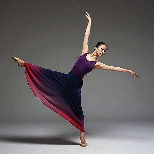 Photograph of a female ballet dancer in a deep purple dress with red gradient, mid-leap, arms gracefully extended, against a gray studio background.