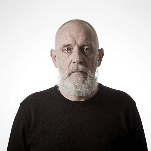 Photograph of an elderly white man with a bald head, white beard, and black sweater, centered against a white background. Serious expression, direct gaze