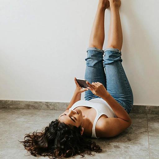 Photograph of a woman with dark curly hair, lying on her back on a tiled floor, wearing a white tank top and blue jeans, holding a