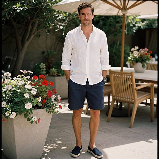 Photograph of a handsome, bearded man in a white button-up shirt, dark shorts, and black slip-on shoes, standing in a sunny garden