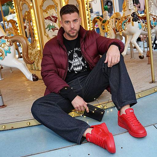 Photograph of a bearded man with short dark hair, sitting on a carousel platform, wearing a maroon puffer jacket, black shirt, dark