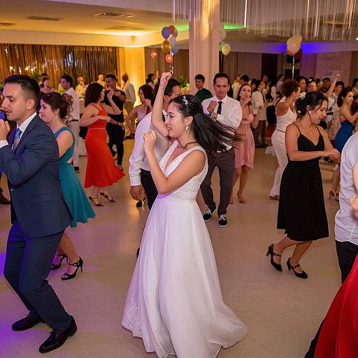 Photograph of a lively wedding reception dance, featuring a bride in a white dress and dark-haired woman dancing in the foreground, surrounded by guests in colorful