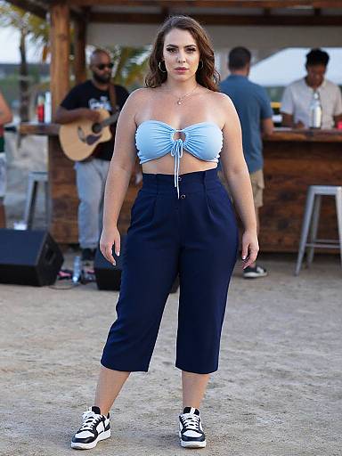 Photograph of a woman with light brown hair, wearing a blue strapless crop top and black high-waisted capri pants, standing outdoors at