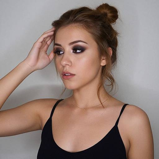 Photograph of a young woman with light skin, brown hair in a bun, wearing black tank top, dramatic smoky eye makeup, gold necklace,