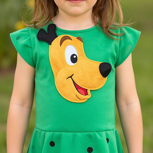 Photograph of a young girl in a green dress with a colorful, cartoon-style dog face on the chest, standing outdoors.