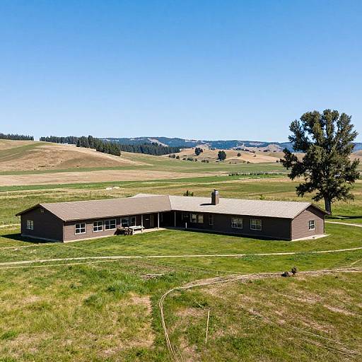 Photograph of a single-story brown house with a pitched roof, set on a grassy hill, surrounded by expansive, sunlit fields and a clear