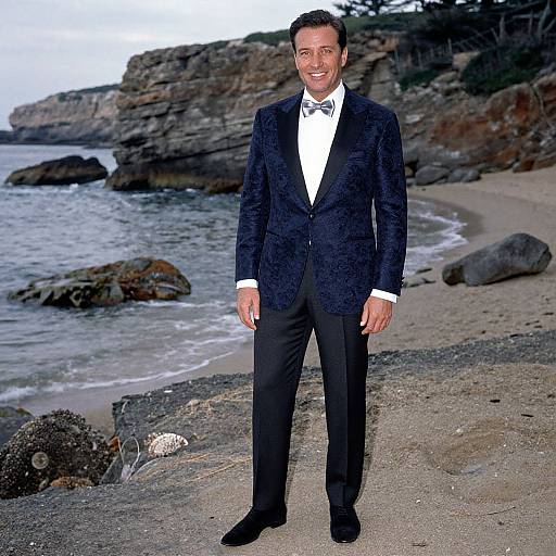 Photograph of a smiling man in a black velvet tuxedo, white shirt, and bow tie standing on a rocky beach with cliffs and ocean in