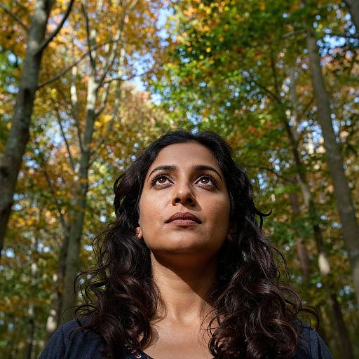 Photograph of a woman with long, wavy black hair, looking upwards, set against a backdrop of autumn trees with yellow and green leaves. She