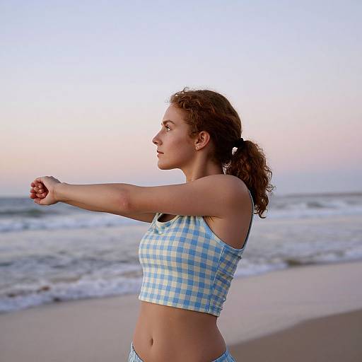 Photograph of a curly-haired woman in a blue and white checkered sports bra stretching on a beach at sunset.