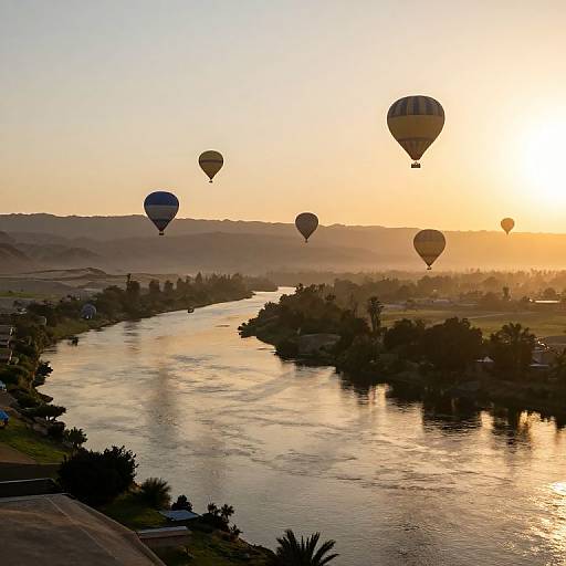 Photograph of a serene sunset over a river with five hot air balloons floating in the sky, casting reflections on the water.