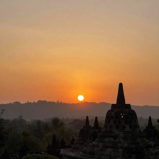 Photograph of a sunset over ancient, silhouetted pagoda-style temples with glowing eyes, set against a gradient orange and yellow sky.