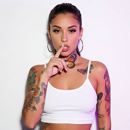 Photograph of tattooed woman with dark hair in a bun, wearing a white crop top, large hoop earrings, finger to lips, colorful butterfly tattoos