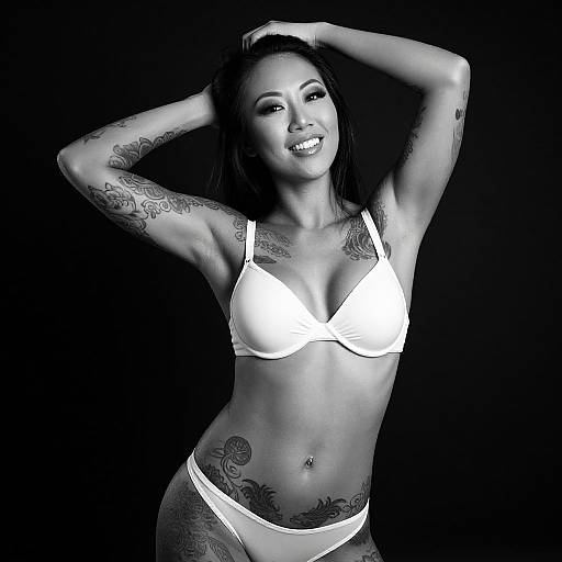 Black-and-white photograph of a smiling, tattooed Asian woman with long hair, wearing white bra and panties, arms raised, dark background.