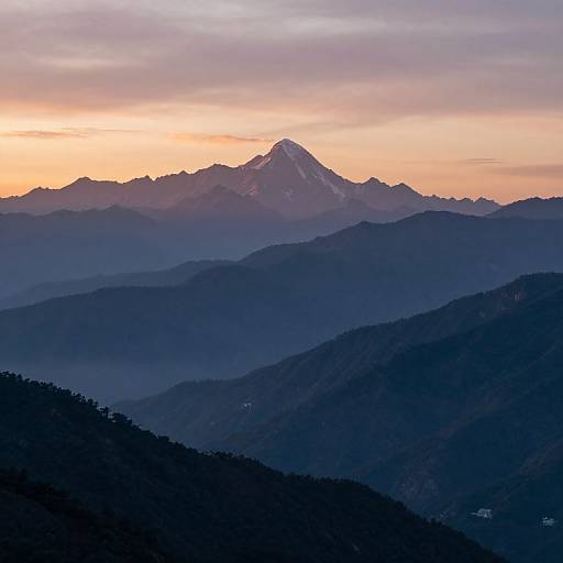 Photograph of a serene mountain landscape at sunset, featuring layered blue and purple mountain silhouettes with a snow-capped peak under a pink and orange