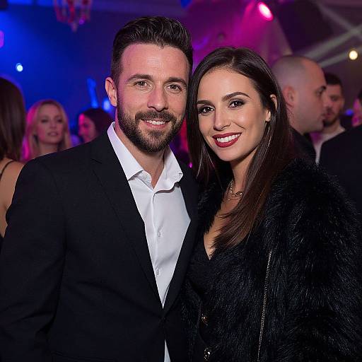 Photograph of a smiling bearded man in a black suit and white shirt, standing next to a woman with long dark hair, wearing a black fur