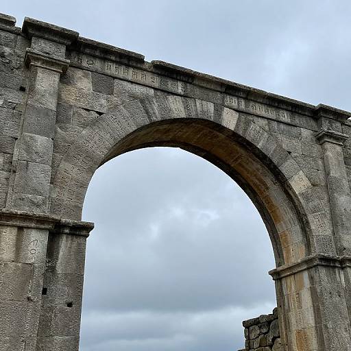 Photograph of a large, ancient stone archway with weathered, gray stones and inscriptions, set against a cloudy sky.